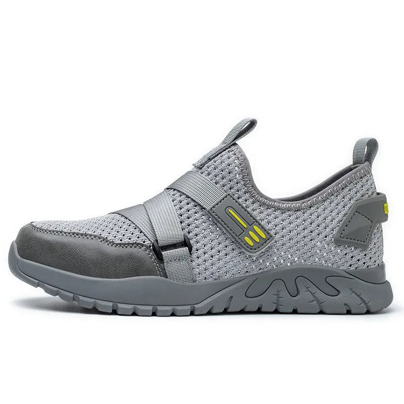 Men's grey safety work shoes with a woven upper and thick rubber sole on a work site background.