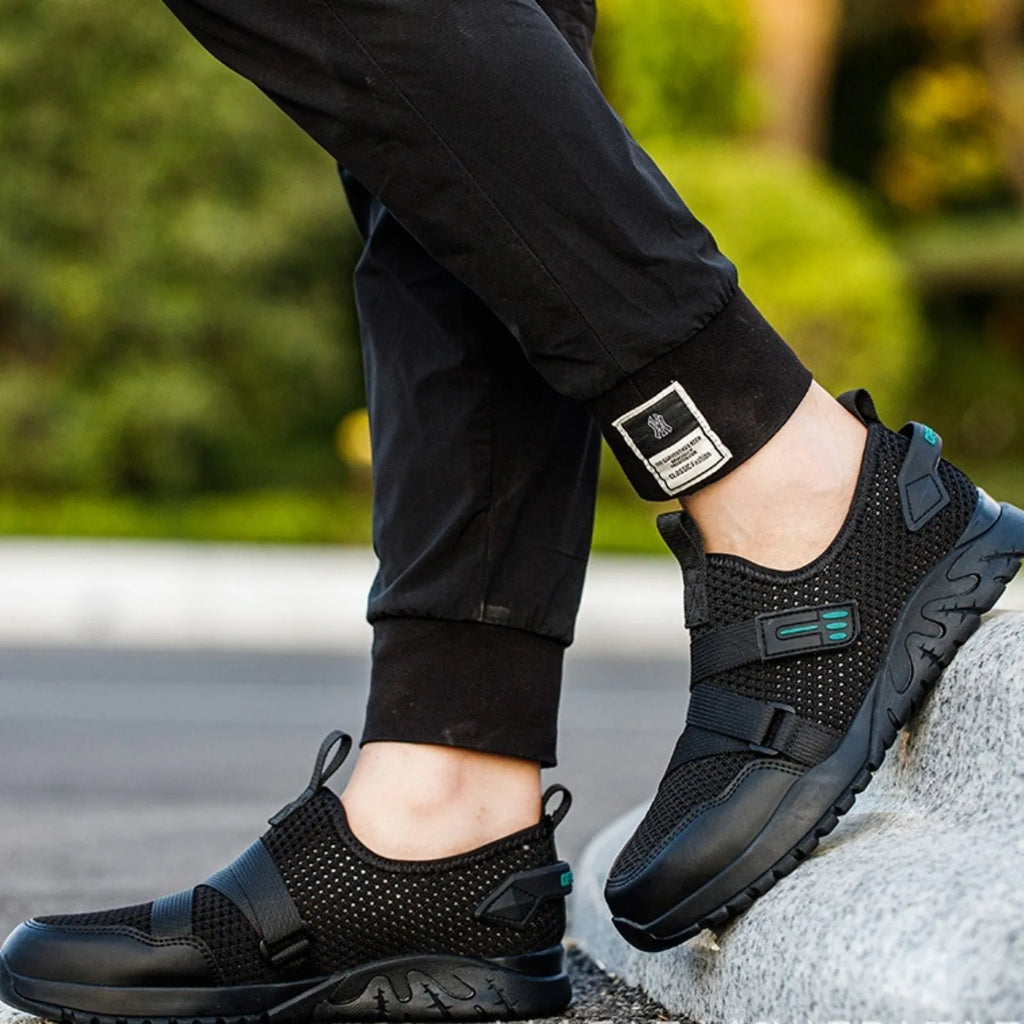 Men's grey safety work shoes with a woven upper and thick rubber sole on a work site background.